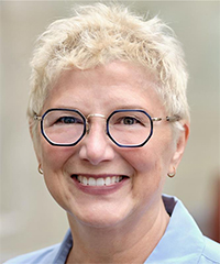 A woman with short, platinum blonde hair and round, silver-framed glasses smiles warmly at the camera. She has light skin, bright eyes, and is wearing a light blue collared shirt. The softly blurred background suggests an outdoor or naturally lit setting.