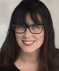 A woman with long dark hair, bangs, and black-framed glasses smiles warmly at the camera. She has light skin, blue eyes, and is wearing a black top. The background is softly blurred, suggesting an indoor setting with natural lighting.