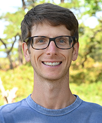 Smiling man with short brown hair, black-rimmed glasses, and a light blue shirt, outdoors with green foliage in the background.