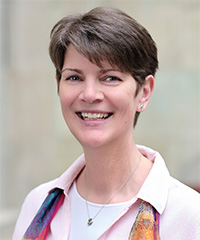 Photo of a woman smiling, with short brown hair, wearing a light pink shirt, a colorful scarf, and small earrings.