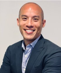 A bald man with a broad smile is wearing a dark suit jacket over a light blue checkered shirt. He is in front of a plain light-colored background.