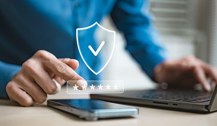 A person in a blue shirt taps a glowing shield-and-checkmark icon above a masked password field, with a laptop and smartphone on a desk in front of them.