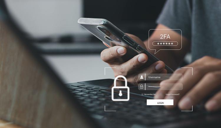A person holding a phone in their right hand while typing on a laptop using their left hand uses authentication to log in to their account